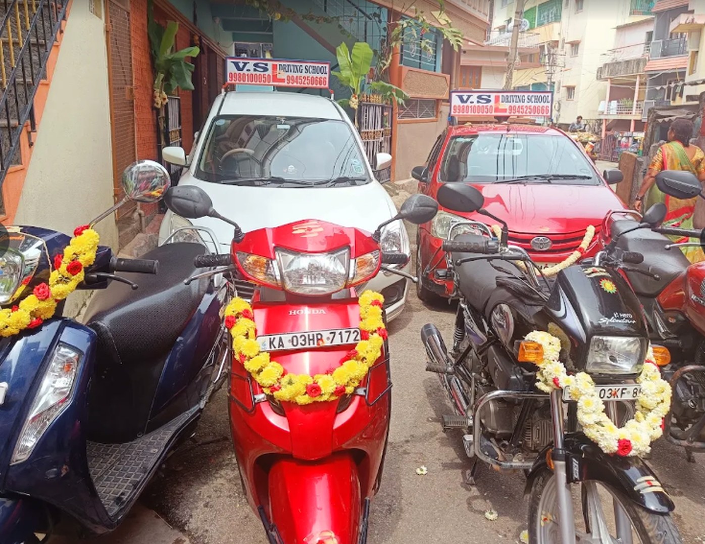 VS Motor's training vehicles parked on the street: two cars with V.S Driving School roof boards, a scooter and a motorcycle, all garlanded